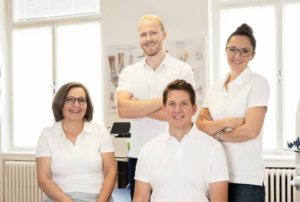 Das Team einer modernen Physiotherapiepraxis in weißer Berufskleidung lächelt im hellen Behandlungsraum.